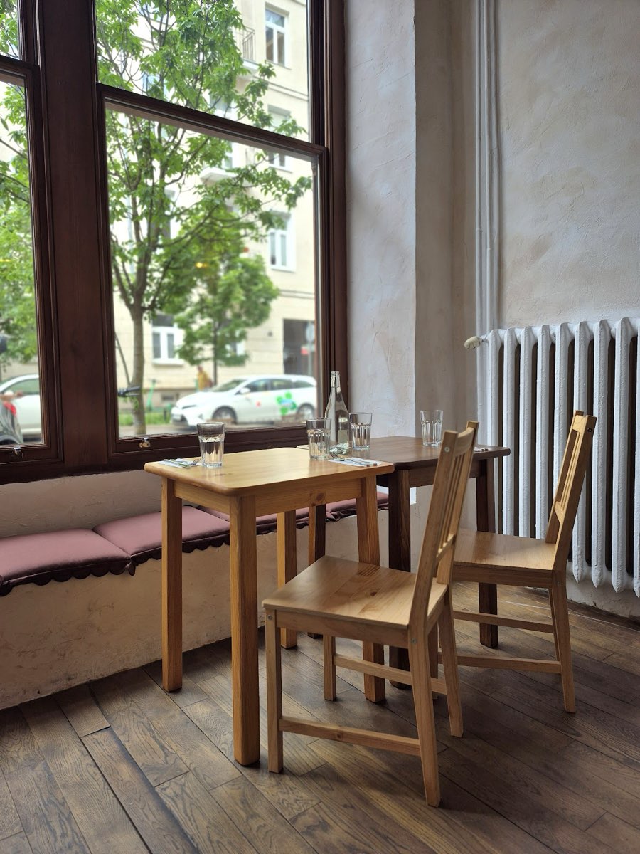 Wnętrze bistro: drewniany stół i krzesła przy dużym oknie kamienicy, różowa ława i tynkowana ściana